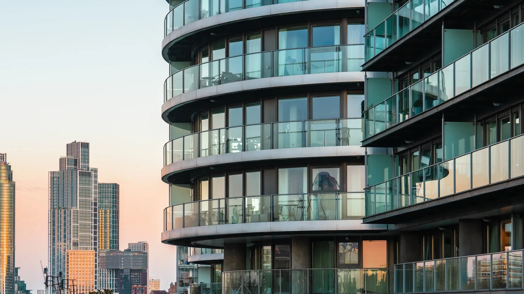 Modern curved apartment building with glass balconies against city skyline at sunset.