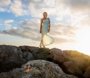 Woman in a flowing dress stands on rocky cliffs at sunset.
