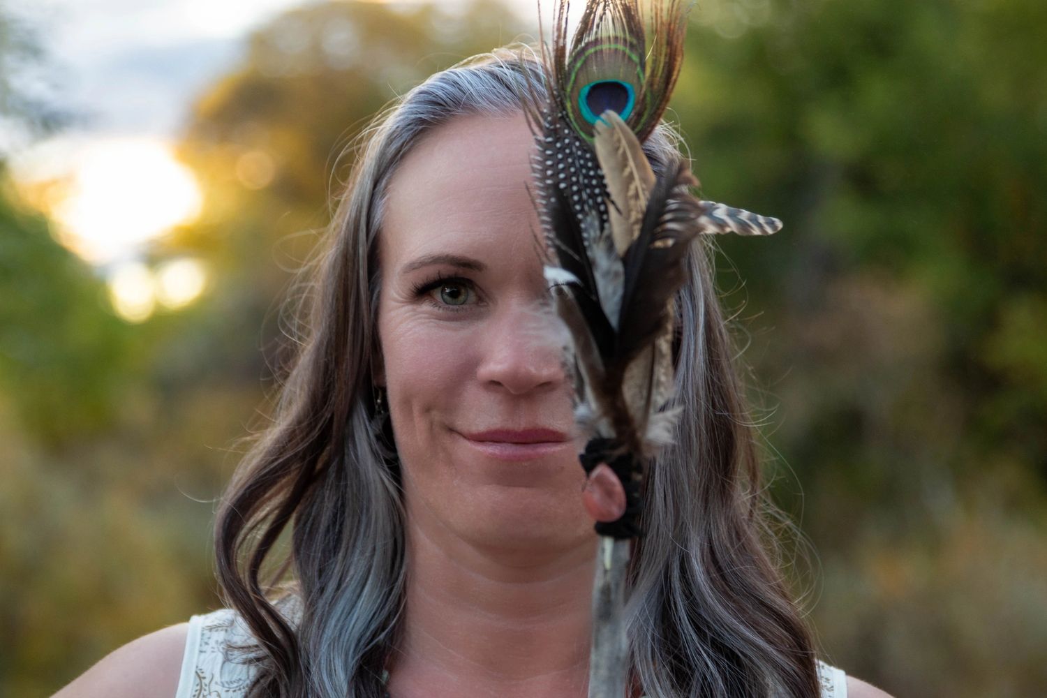 Woman holding a stick decorated with feathers covering half her face outdoors.