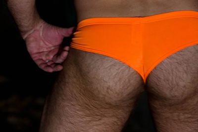 Man wearing bright orange briefs standing with hands on hips in the dark.