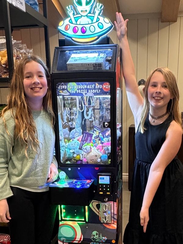 Two girls smiling by a claw machine with snacks on shelves behind.