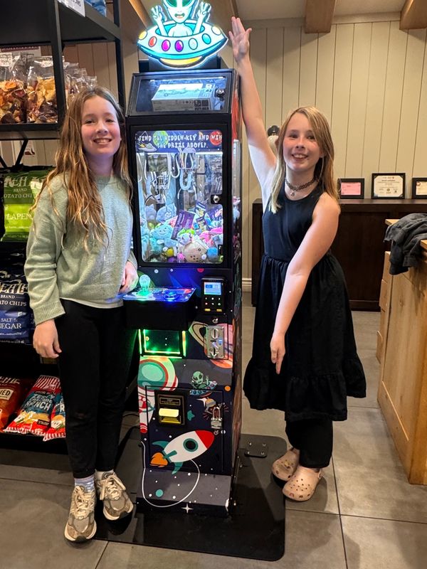 Two girls smiling by a claw machine with snacks on shelves behind.