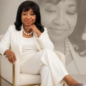 Confident woman in white suit sitting thoughtfully on a chair with a serene expression.