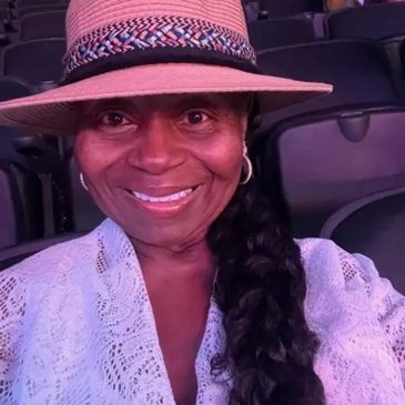 Smiling woman wearing a hat and white lace top in a seated area.
