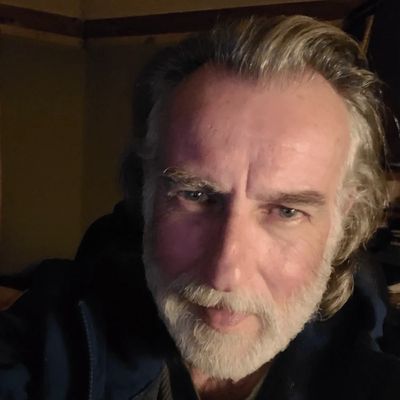 Close-up of a bearded man with gray hair in dim lighting.