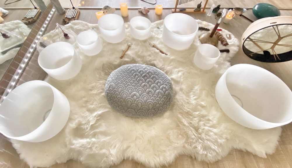 Looking down on white crystal singing bowls of different sizes set in a circle on a white sheepskin rug around a grey patterned floor cushion, with a frame drum to the right
