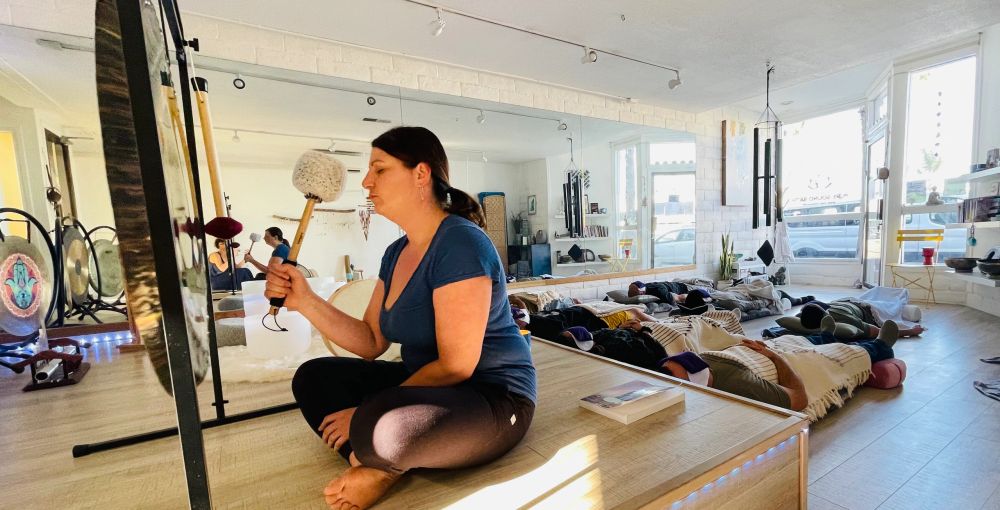The studio during a session, a practitioner seated on a low wooden platform holding a padded gong mallet beside a large gong, while participants lie under blankets on the floor in front of a mirrored wall and the storefront windows