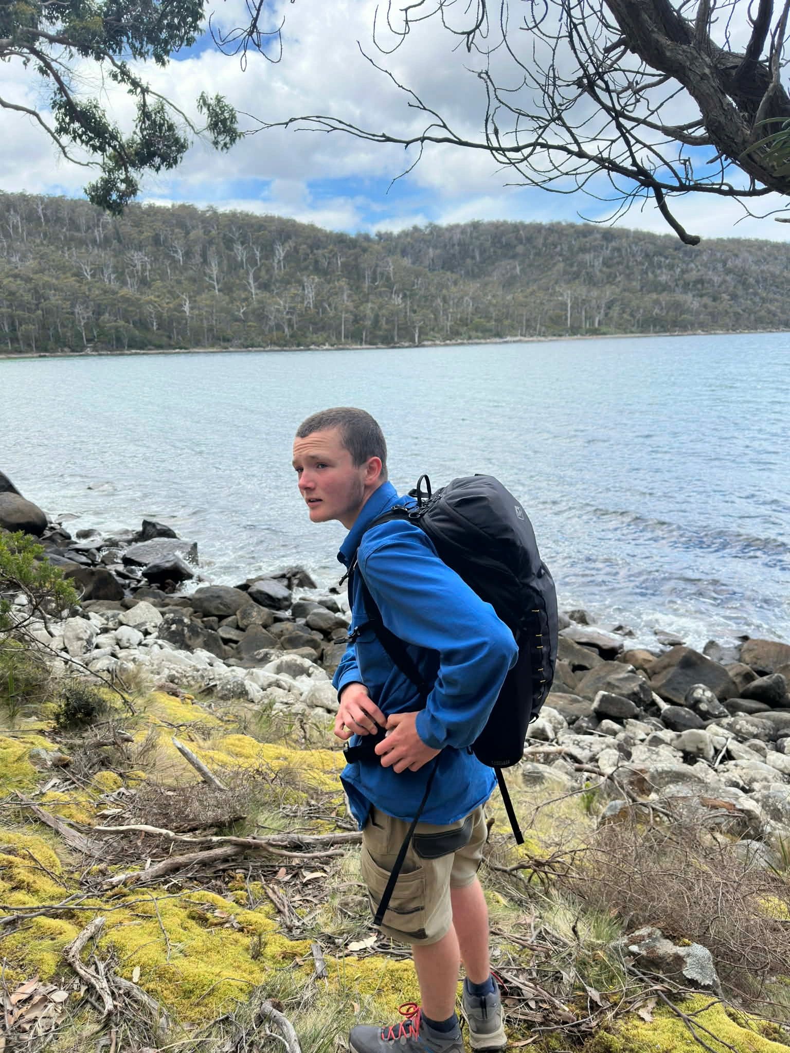 James Repine hiking along a waterfront trail with a forest in the background and blue sky.