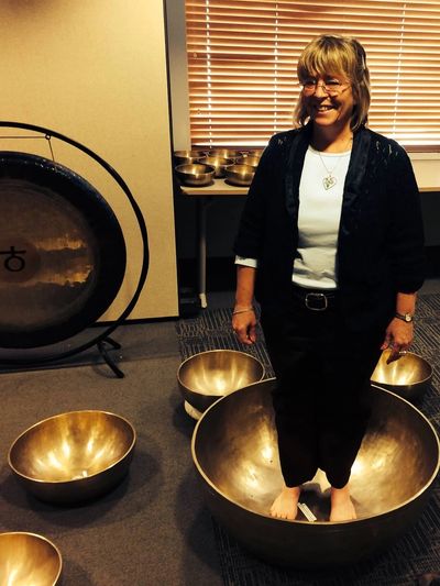 Woman standing barefoot inside a large singing bowl, surrounded by other bowls and a gong.