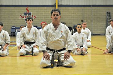 Two martial arts instructors sitting with students on a dojo floor.