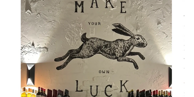 Wall art of a leaping rabbit with the phrase 'Make Your Own Luck' above wine bottles.