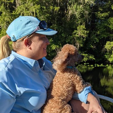 Woman in blue holding a curly-haired dog near a river.