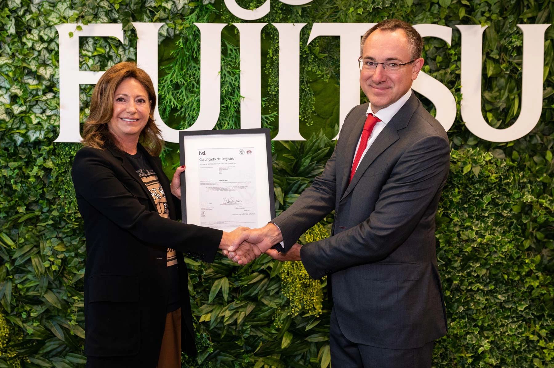 Two professionals shake hands as one holds a certificate against a green leafy backdrop.