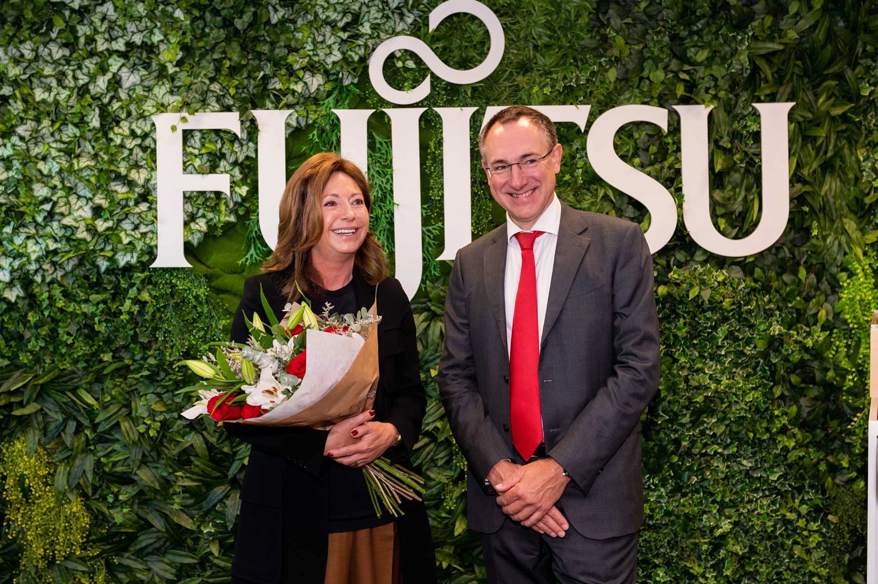 Two people smiling in front of a green leafy backdrop with Fujitsu logo, one holding a bouquet.