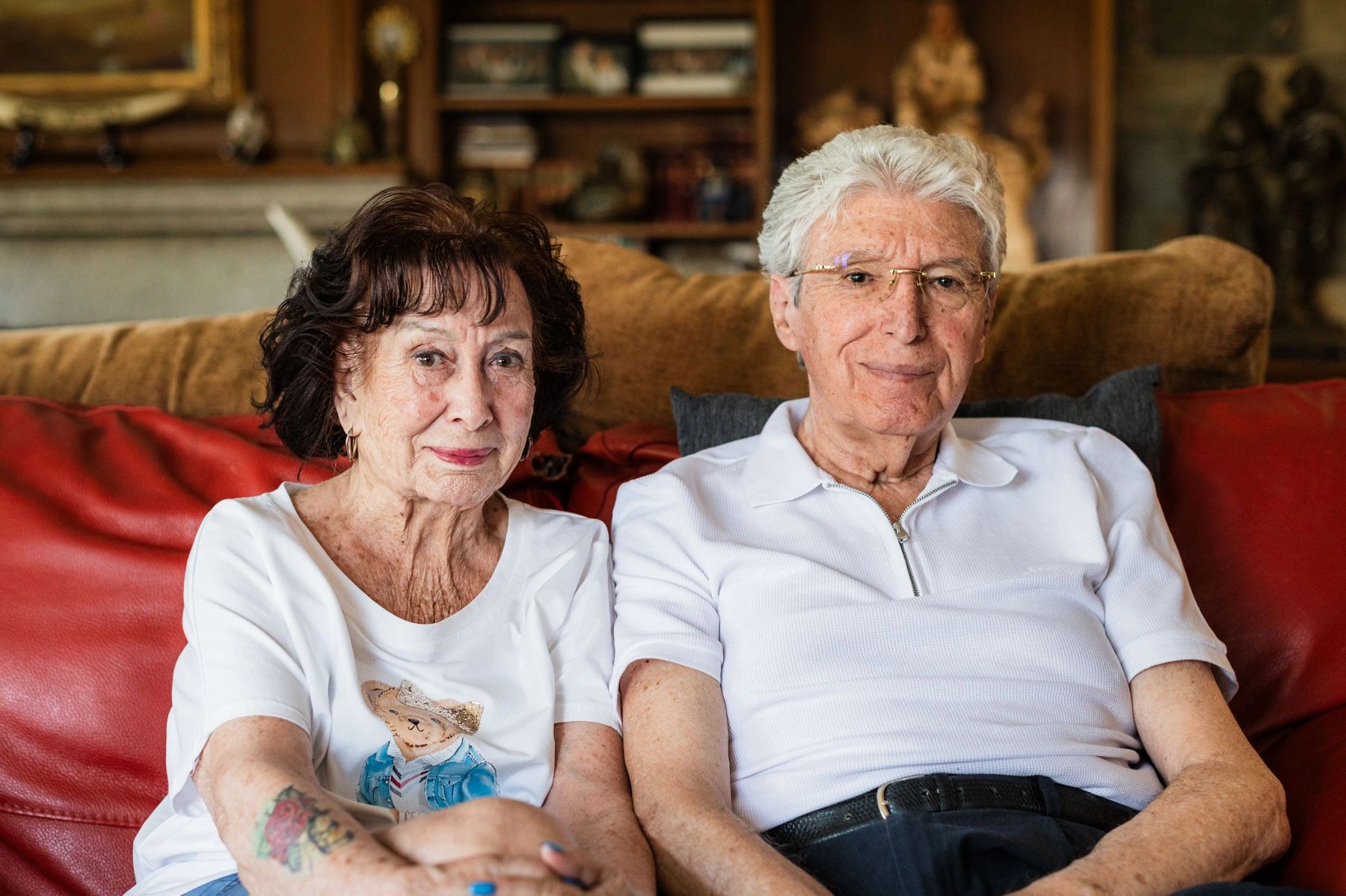 Elderly couple sitting together on a red couch, smiling gently.
