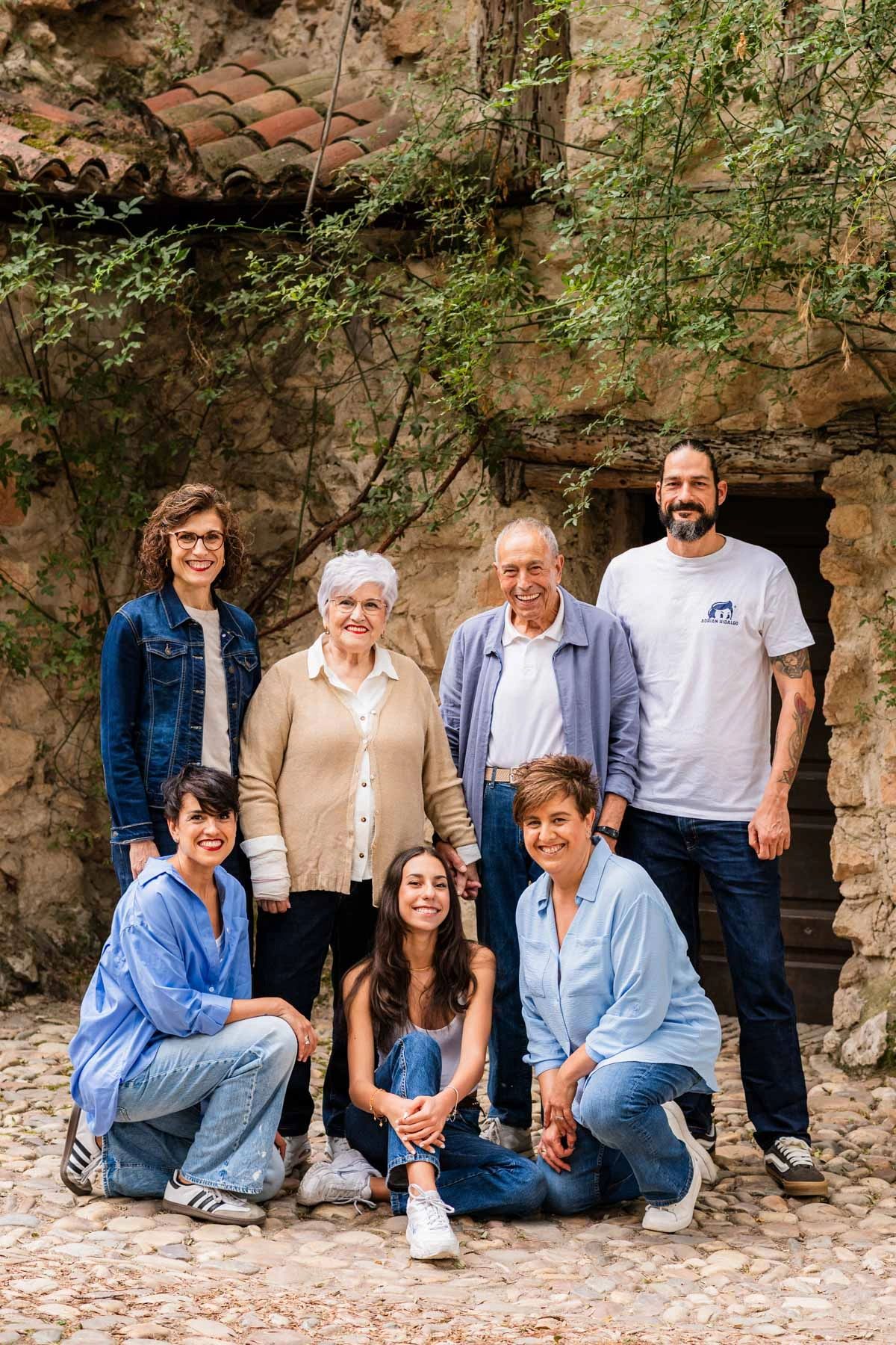 A multi-generational family posing outdoors in casual attire with a rustic stone wall background.