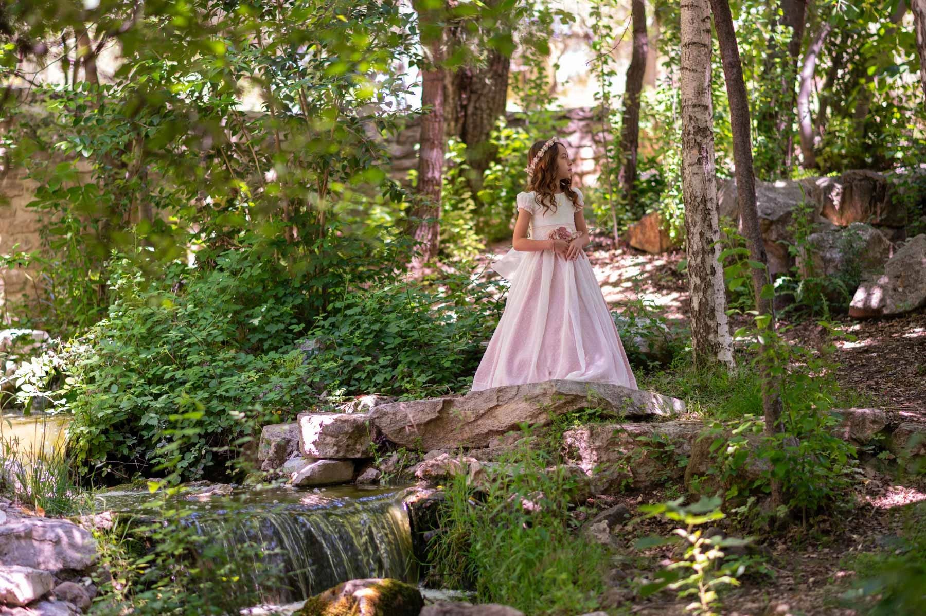 A girl in a pink dress stands on a stone bridge in a lush forest.