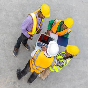 Construction workers in helmets and safety vests reviewing plans on a laptop.