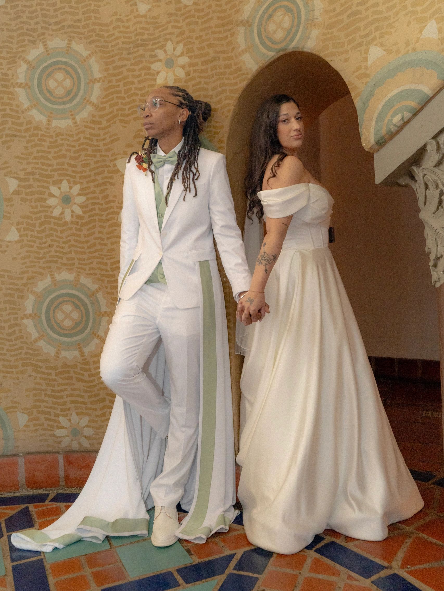 Couple in wedding attire holding hands, posing against a decorative wall.