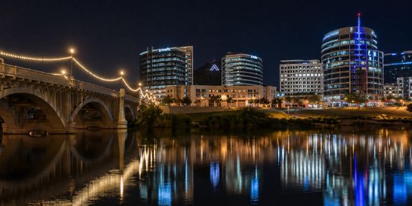 City skyline and bridge lit up at night, reflecting on water.