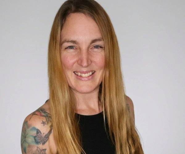 Smiling woman with long blonde hair and tattoos, wearing a black sleeveless top.