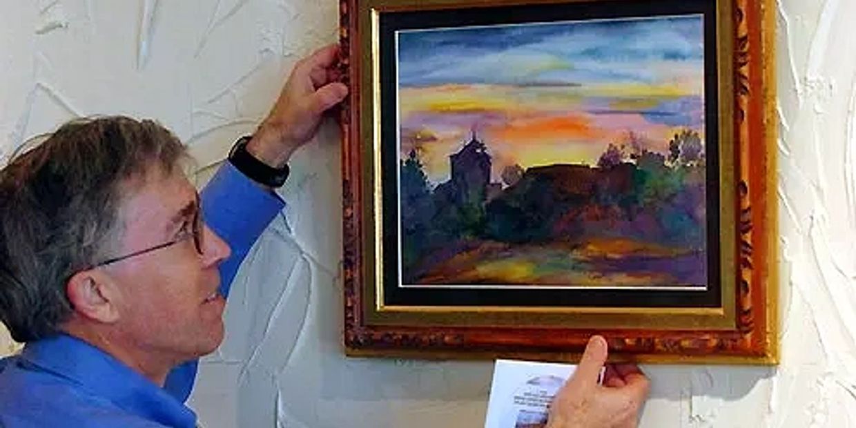 Man hanging a framed sunset painting on a textured wall.