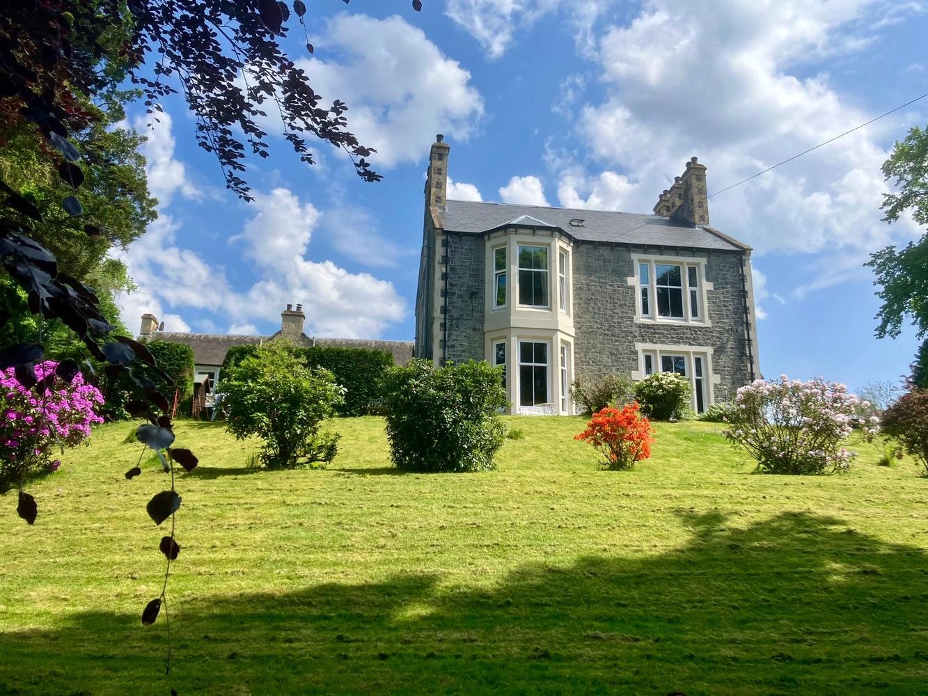 A large stone house with a manicured garden under a partly cloudy blue sky. above and beyond moffat