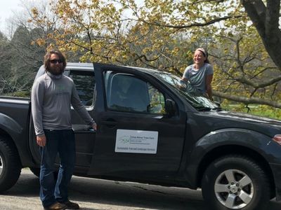 Two people posing with a black pickup truck under autumn trees.