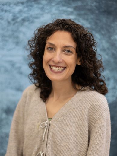Smiling woman with curly hair in a beige sweater against a blue background.