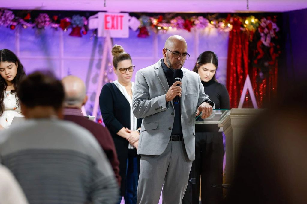 A man in a gray suit leads a prayer with others standing behind him in a decorated room.