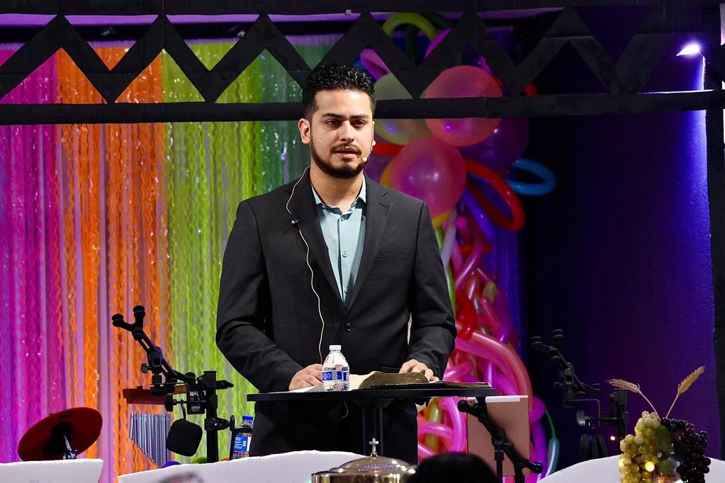 A man in a black suit speaks on stage with colorful balloon decorations behind him.