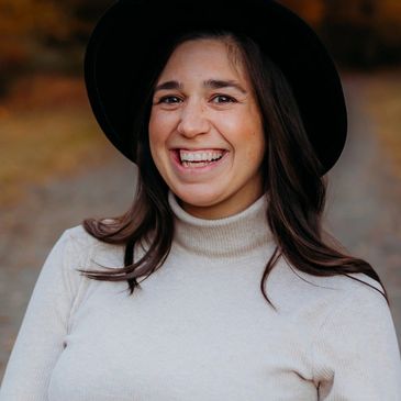 Woman smiling outdoors wearing a black hat and beige turtleneck.