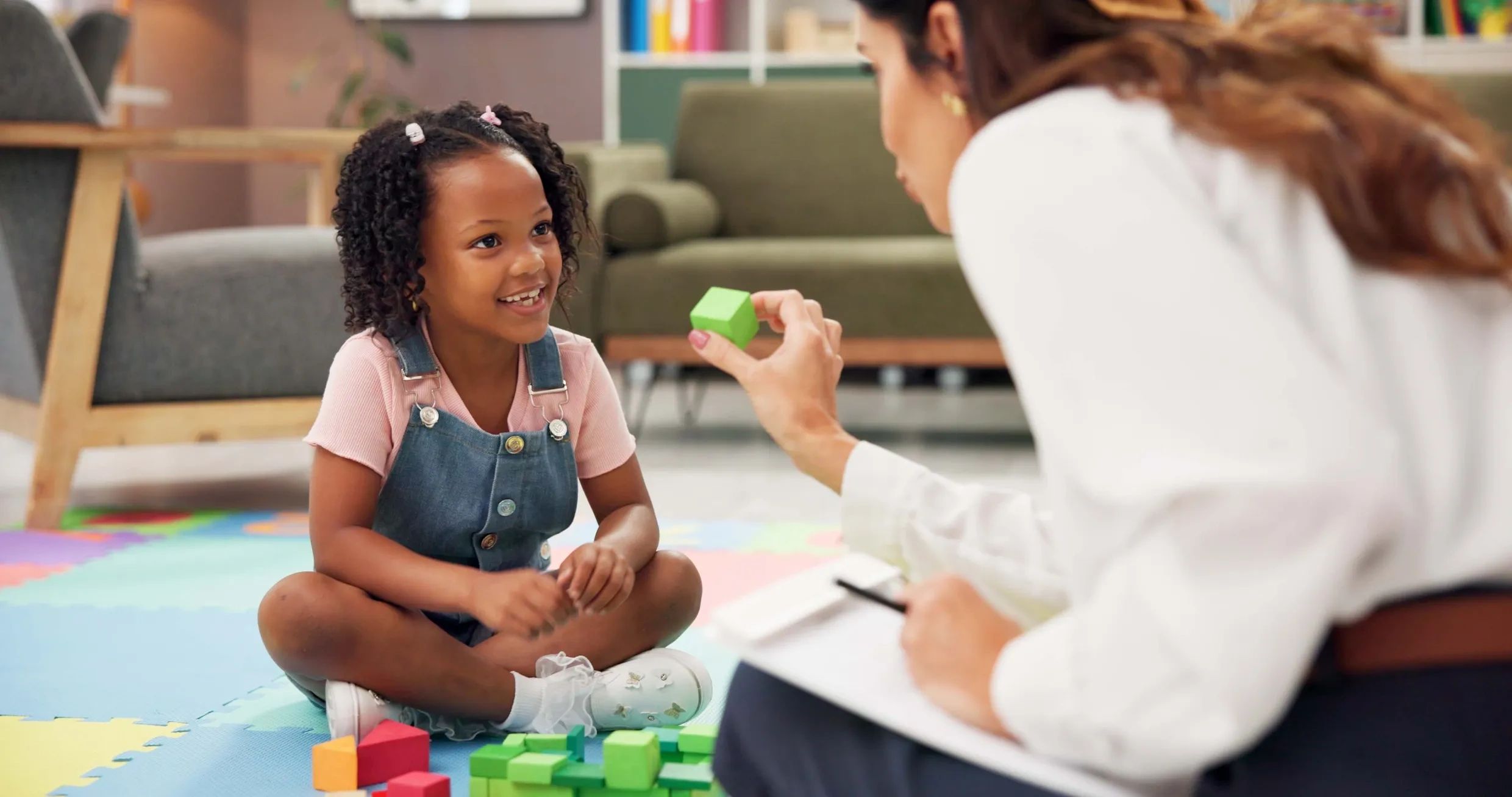 A young girl smiling while playing with colorful blocks and interacting with an adult.