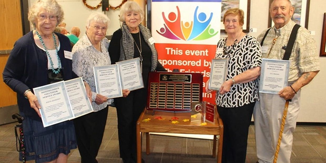 Five senior volunteers for the Friends of the Library and Museum of LIttleton who were honored for their service. Three women on the left and one man on the right stand in front of the Welcome Friends banner at the Bemis Public Library in Littleton. 