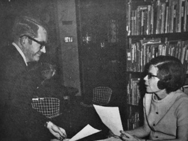 A man smiles as he hands over paperwork to a young woman sitting in front of a bookshelf. 