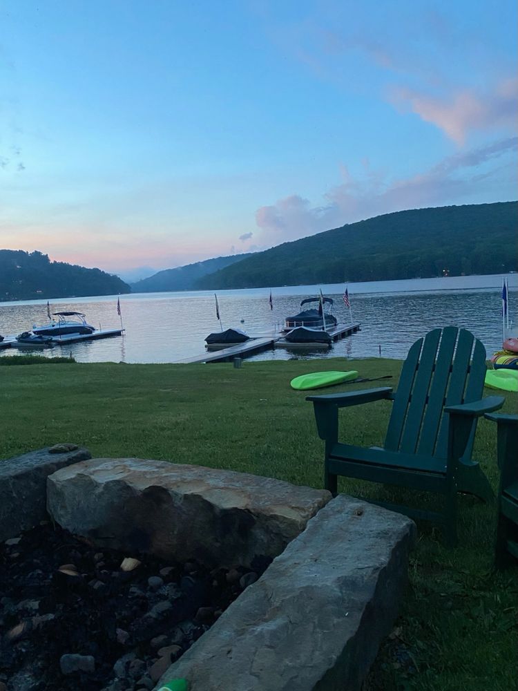 Peaceful lakeside view with boats, Adirondack chairs, and a stone fire pit at dusk.