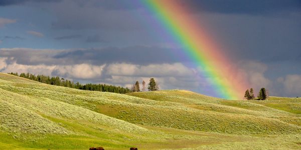 A vibrant rainbow stretches over rolling green hills under a cloudy sky.