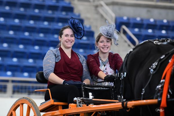 Two women in fancy hats smiling on a horse-drawn carriage indoors.