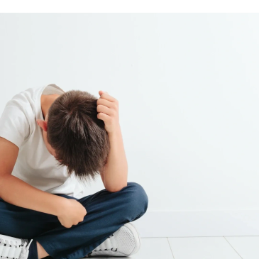 A boy sitting on the floor looking down, holding his head.