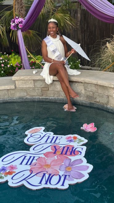 Woman in white dress and tiara sitting by pool at celebration.
