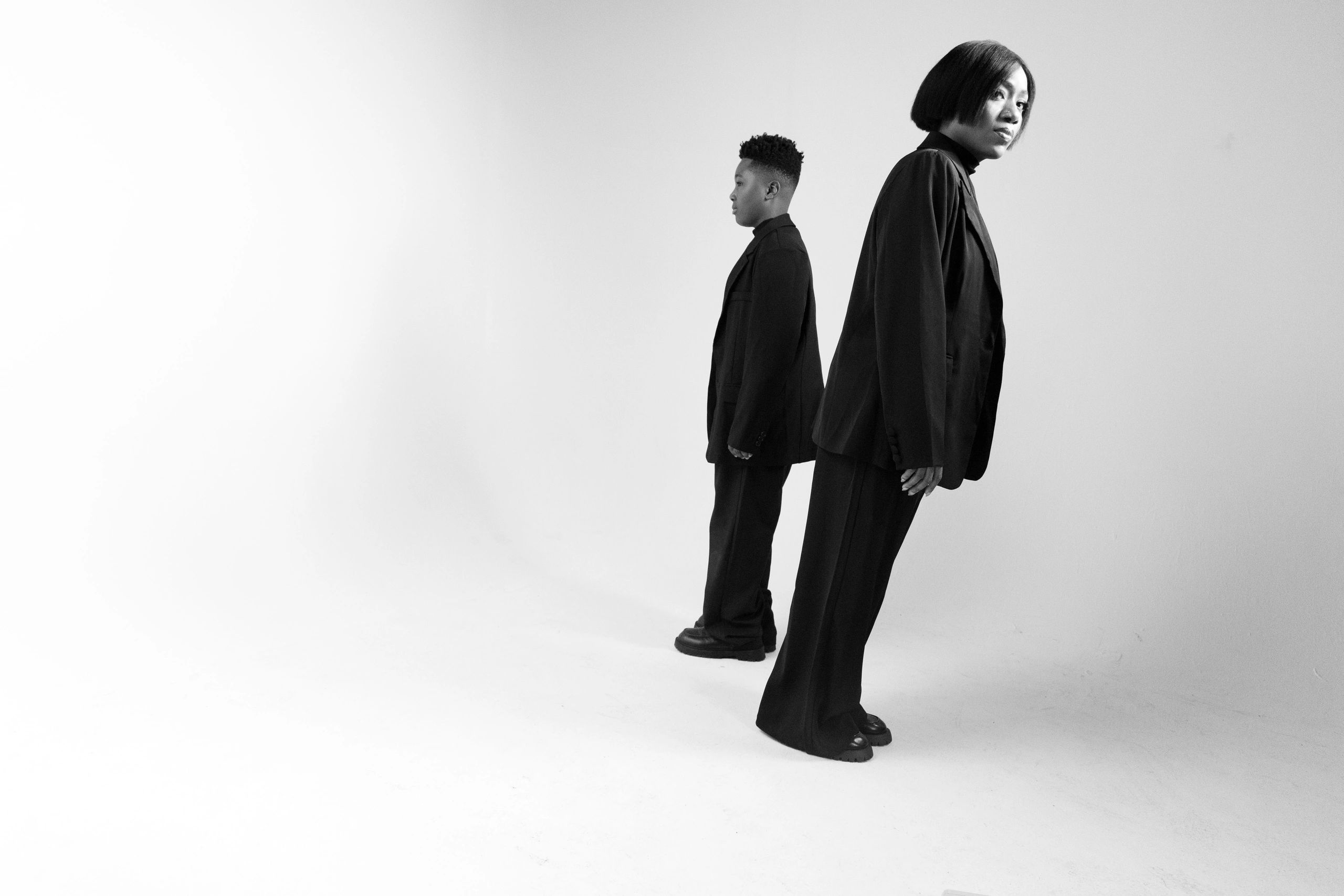 Two people in black outfits posing back-to-back in a studio.
