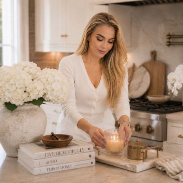 Woman lighting a candle in a cozy kitchen setting with flowers and books.