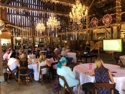 People attending a presentation in a rustic barn decorated with string lights and chandeliers.