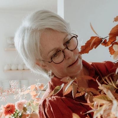 Elderly woman with glasses arranging autumn leaves indoors.