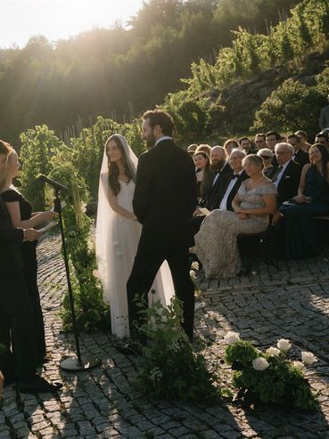 A couple stands at an outdoor wedding ceremony surrounded by guests and greenery.