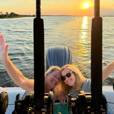 Two women enjoying a boat ride at sunset, smiling and waving.