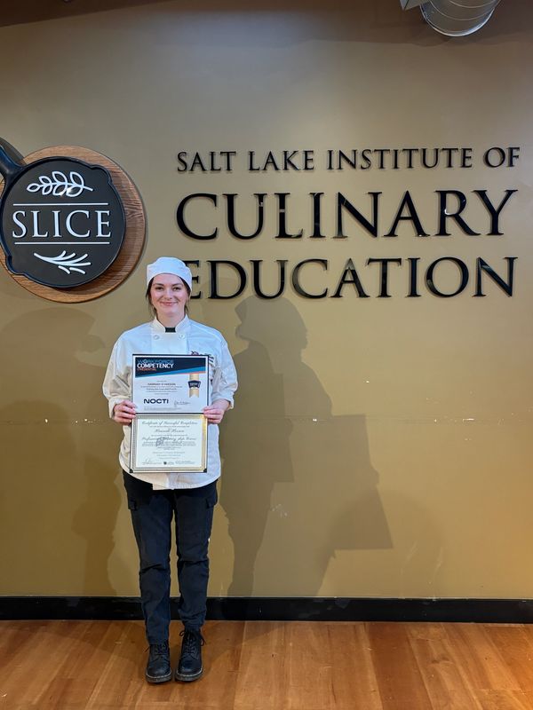 A woman chef proudly holding certificates in front of a culinary education sign.