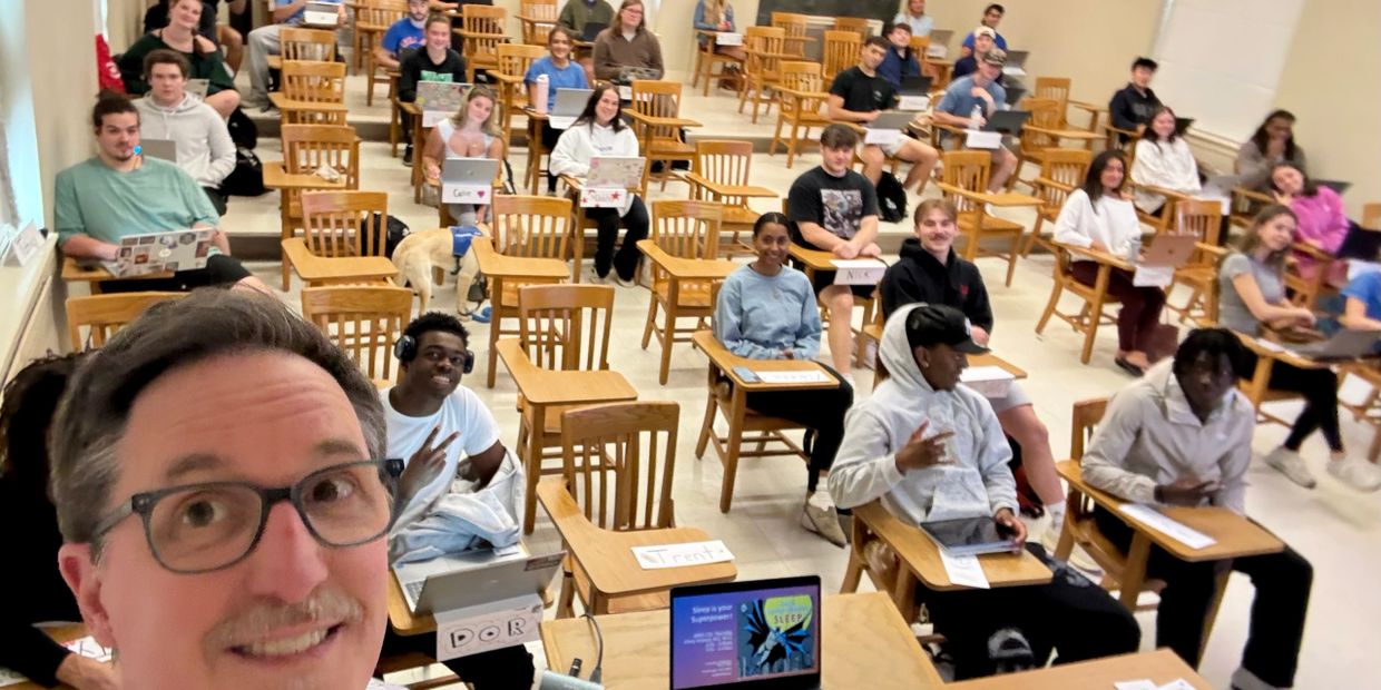 A teacher taking a selfie with students seated in a classroom with laptops and name tags.
