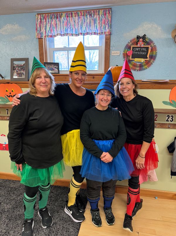 Four women dressed as colorful crayons pose happily in a classroom.