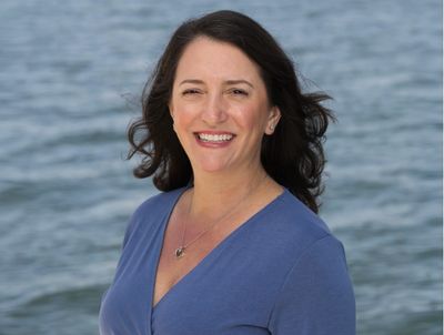 Smiling woman in blue top by the water with wind-blown hair.